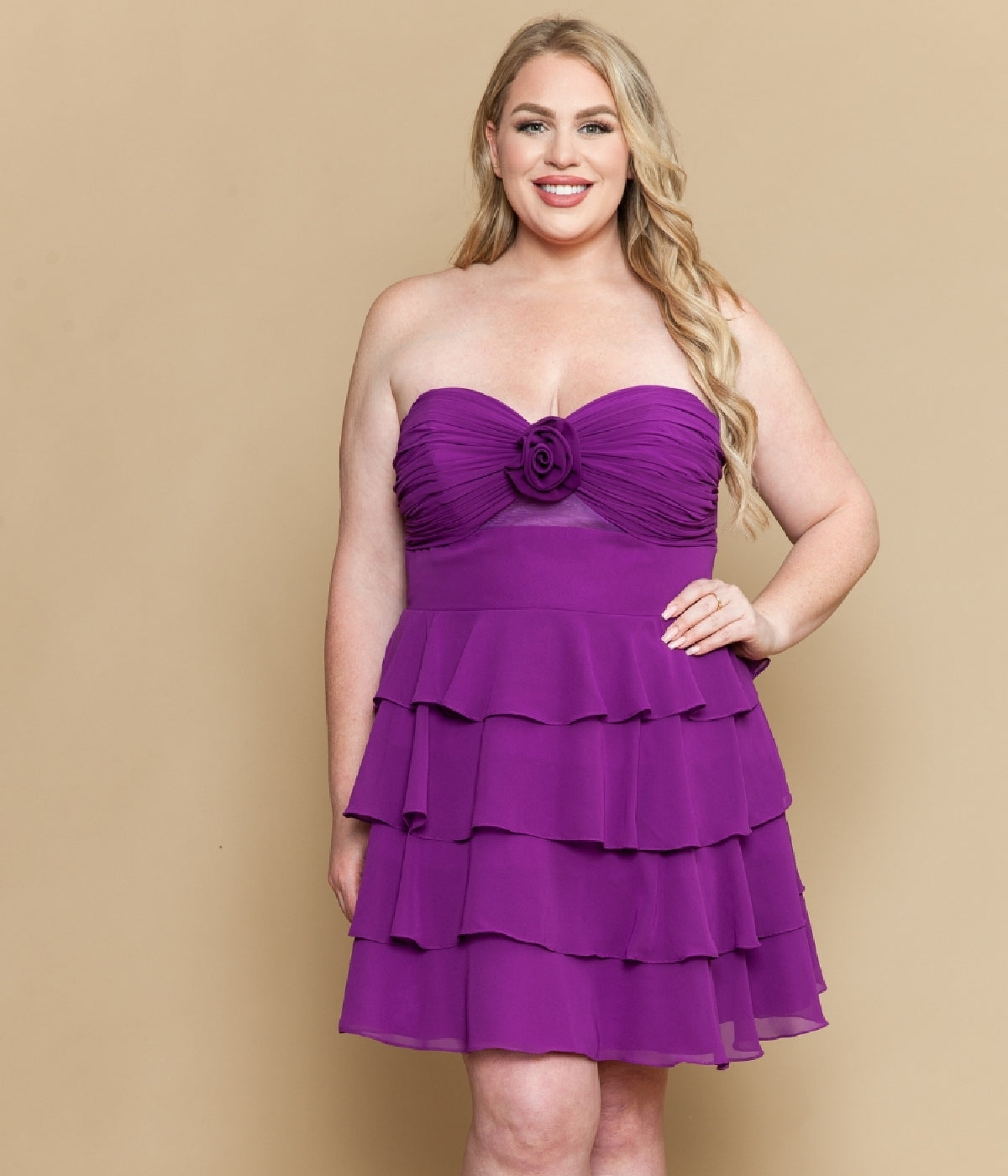 A woman models the Plus-Size Short Ruffle Party Dress: Poly USA W1218, featuring purple tiers and a floral bodice accent, standing against a plain beige background and smiling at the camera.